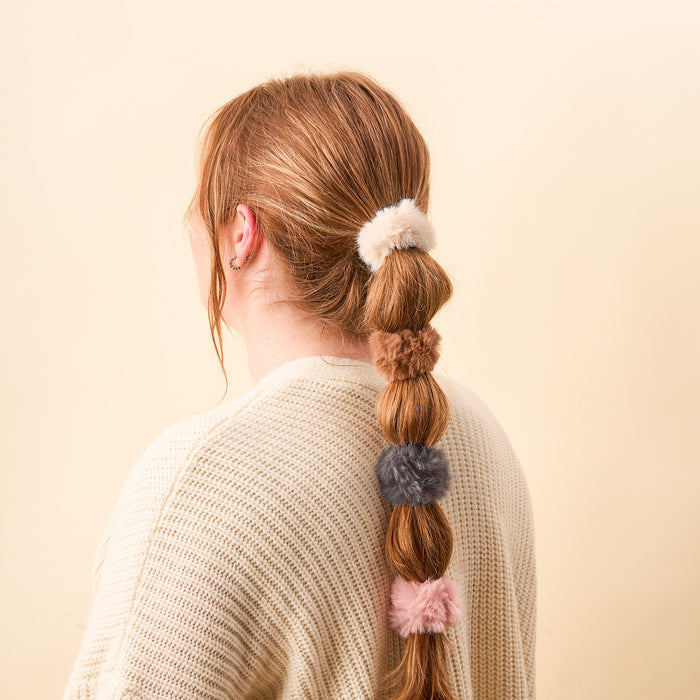 A person with long reddish hair, tied in sections with four Bliss Faux Fur Scrunchies by The Darling Effect in white, brown, gray, and pink, pairs the luxury accessories with a cream knit sweater against a pale beige background.