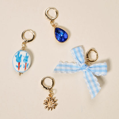 Four small gold keychains from The Darling Effect's Blue Belle The Ultimate Charm Pack—featuring a blue gem, a light blue painted oval bead, a gold sunburst charm, and a blue gingham ribbon bow—are arranged on a beige background.