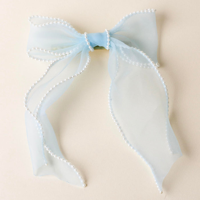 The-darling-effect Blue Sheer Pearl Hair Bow features a light blue organza bow with pearl-beaded edges, gold barrette clip, and long tails—an elegant hair accessory for any occasion. Displayed on a cream background.