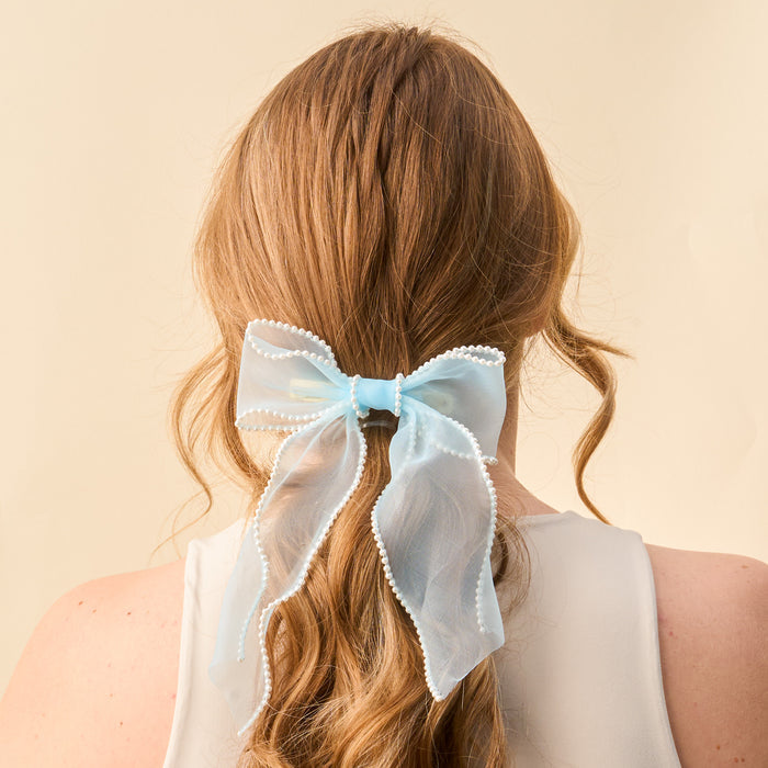 A woman with long, wavy, light brown hair wears the-darling-effect's Blue Sheer Pearl Hair Bow with decorative trim, tied at the back. She is in a sleeveless white top against a beige background.