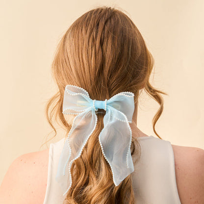A woman with long, wavy, light brown hair wears the-darling-effect's Blue Sheer Pearl Hair Bow with decorative trim, tied at the back. She is in a sleeveless white top against a beige background.