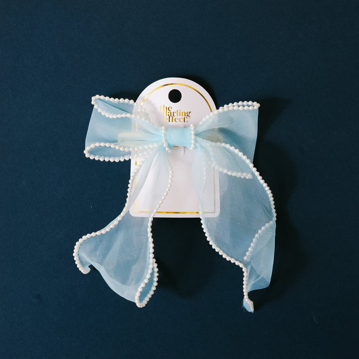 The Blue Sheer Pearl Hair Bow by the-darling-effect features a sheer light blue bow with white trim on a gold barrette, displayed on a white card with gold text against a dark blue background.