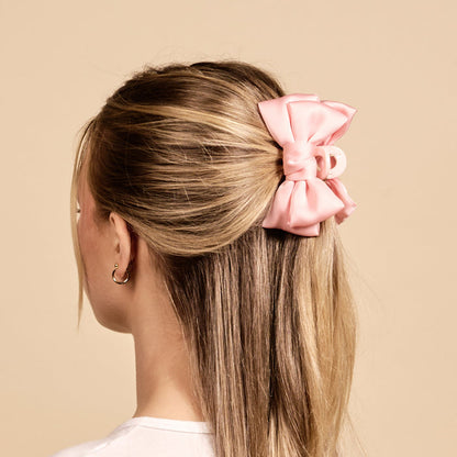 A person with blonde hair wears The Darling Effect's Blush Mini Satin Bow Claw Clip—a large, pale pink bow with a satin finish and a blush claw backing.