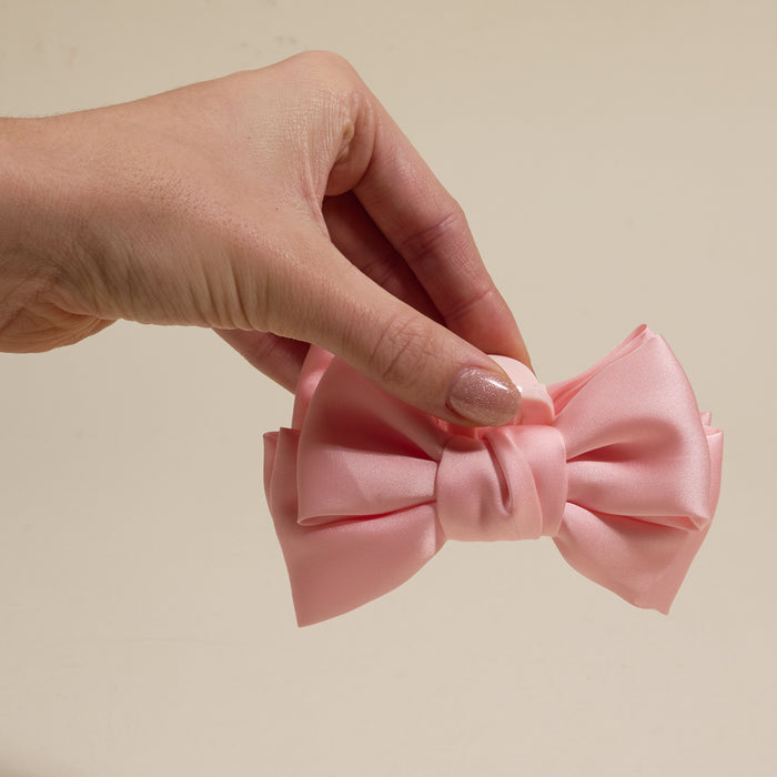 A hand displays The Darling Effect’s Blush Mini Satin Bow Claw Clip—a chic light pink satin hair accessory—against a plain beige background.