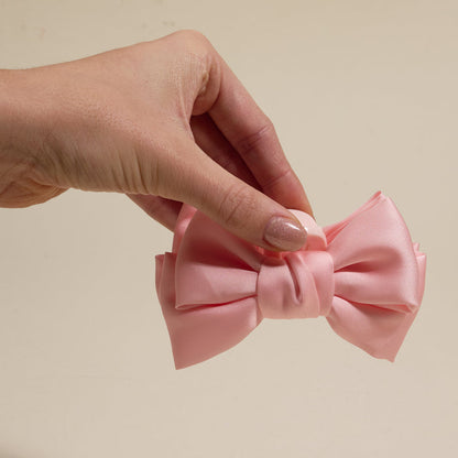 A hand displays The Darling Effect’s Blush Mini Satin Bow Claw Clip—a chic light pink satin hair accessory—against a plain beige background.