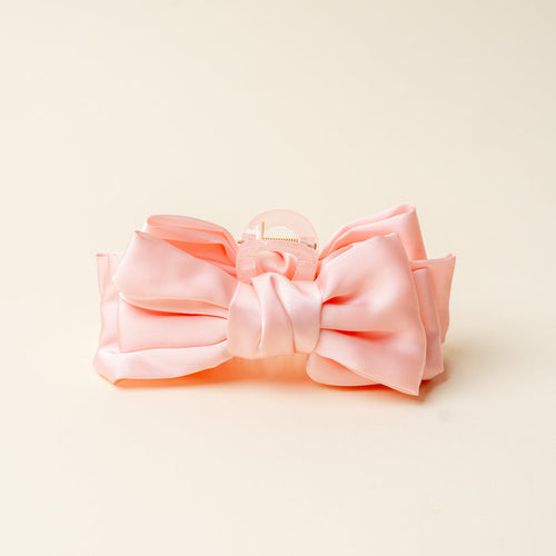 The Darling Effect’s Blush Satin Bow Claw Clip, an elegant hair accessory with a clear plastic clasp, is displayed on a light yellow background.