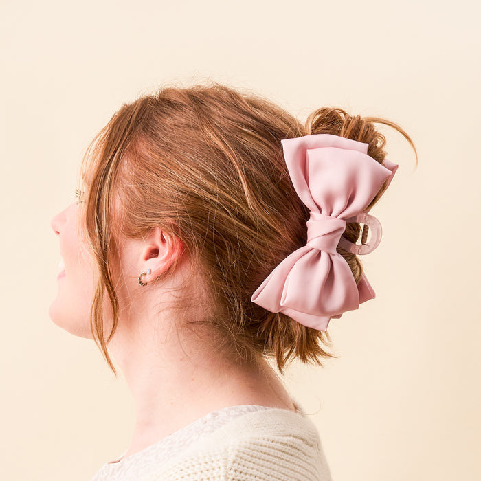 A woman with light brown hair in a loose bun models The Darling Effect’s Blush Satin Bow Claw Clip, showing off the large pink satin accessory from behind and the side against a light beige background.