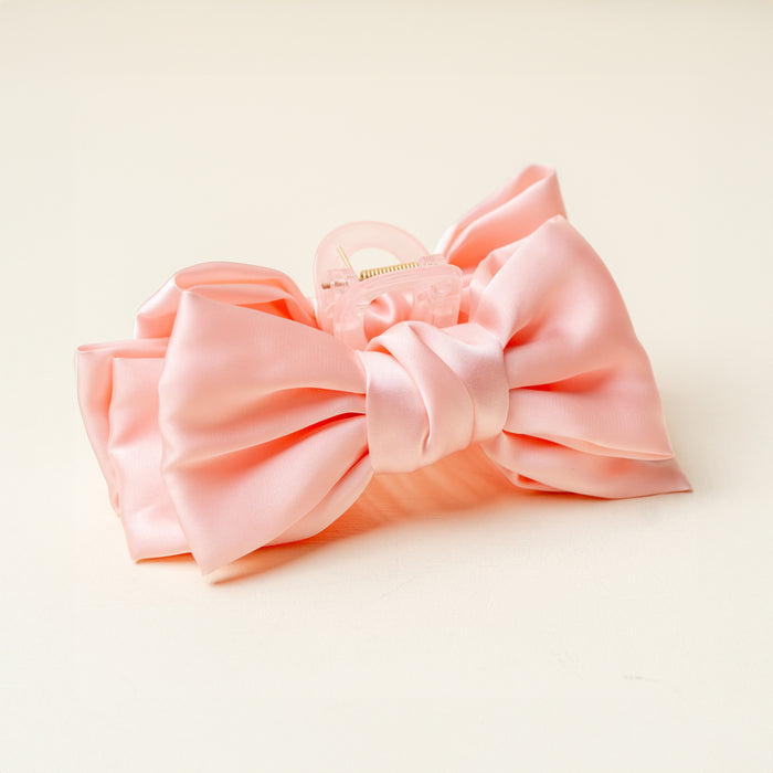 The Darling Effect’s Blush Satin Bow Claw Clip is shown on a cream background. This elegant hair accessory features a light pink satin bow on a clear claw, perfect for adding charm to any look.