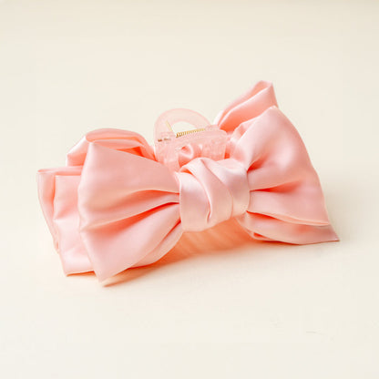 The Darling Effect’s Blush Satin Bow Claw Clip is shown on a cream background. This elegant hair accessory features a light pink satin bow on a clear claw, perfect for adding charm to any look.