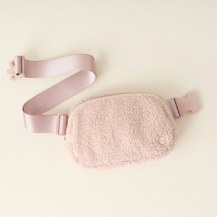 The Darling Effect Blush Sherpa Belt Bag features soft polyester sherpa fabric in light pink, with a matching adjustable strap and plastic buckle, displayed on a cream-colored surface.