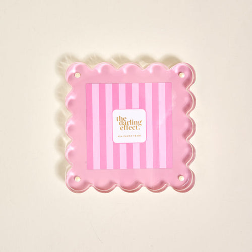 The Darling Effect’s Blush Square Scalloped Acrylic Picture Frame features pink scalloped edges, a striped background, and corner holes for mounting. The modern frame showcases “the darling effect” at its center on a light surface.