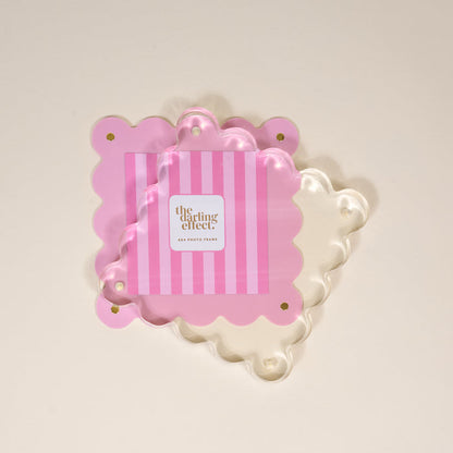 Two Blush Square Scalloped Acrylic Picture Frames by The Darling Effect, one pink stripe and one clear, overlap. The pink magnetized frame displays “darling effect” in gold at the center, adding a charming modern touch.