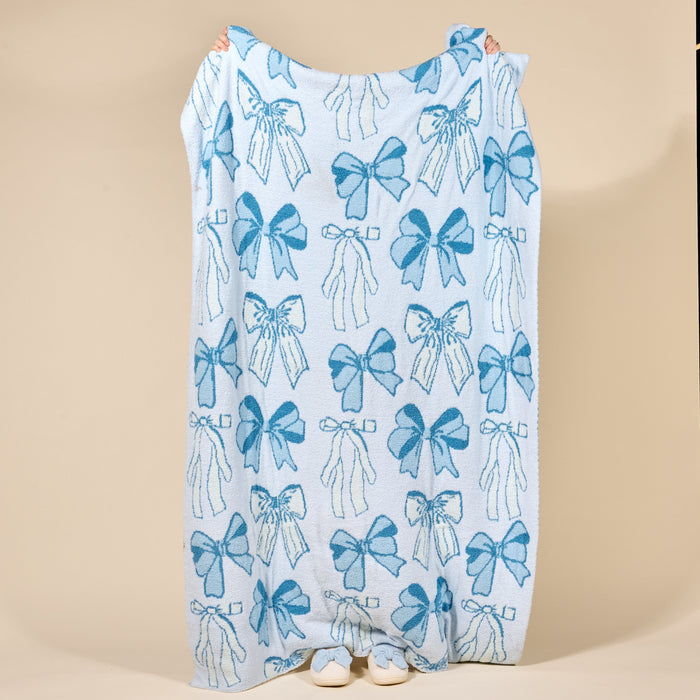 A person holds up the-darling-effect’s Blushing Bows Blue Cozy Dreams Luxe Blanket, featuring blue bow designs. Only their hands and white shoes show behind the ultra-soft blanket against a beige background.