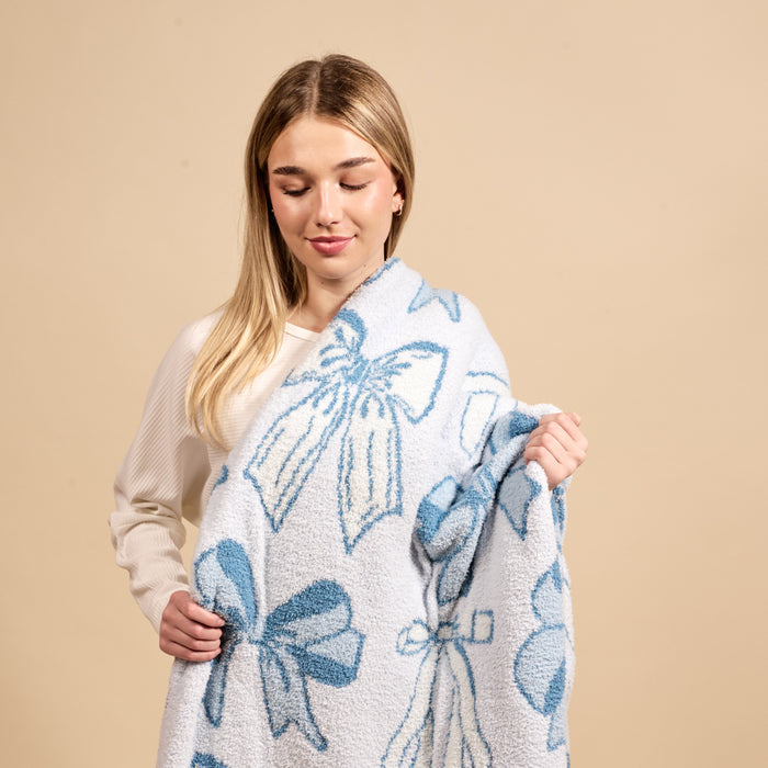A young woman with long blonde hair in a cream sweater holds the-darling-effect’s Blushing Bows Blue Cozy Dreams Luxe Blanket, featuring large blue bow patterns on a light blue and beige background.