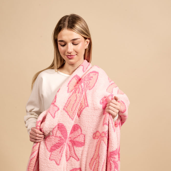 A young woman with straight blonde hair and a white top gently smiles down while holding the-darling-effect’s Blushing Bows Pink Cozy Dreams Luxe Blanket—an ultra-soft throw featuring large pink bows—against a plain beige background.