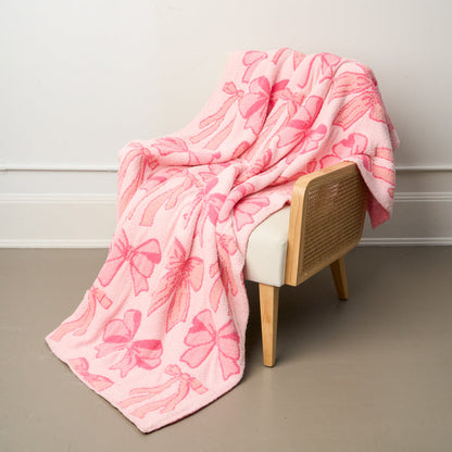 The-darling-effect's Blushing Bows Pink Cozy Dreams Luxe Blanket, made from ultra-soft polyester microfiber with pink bow patterns, is draped over a light wood and cane chair with a white cushion against a beige floor and white wall.