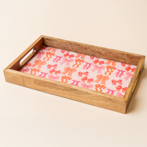 The-darling-effect's Blushing Bows Rectangle Wood Enamel Tray features handles, a pink base, and an orange and pink bow pattern on a light beige background.