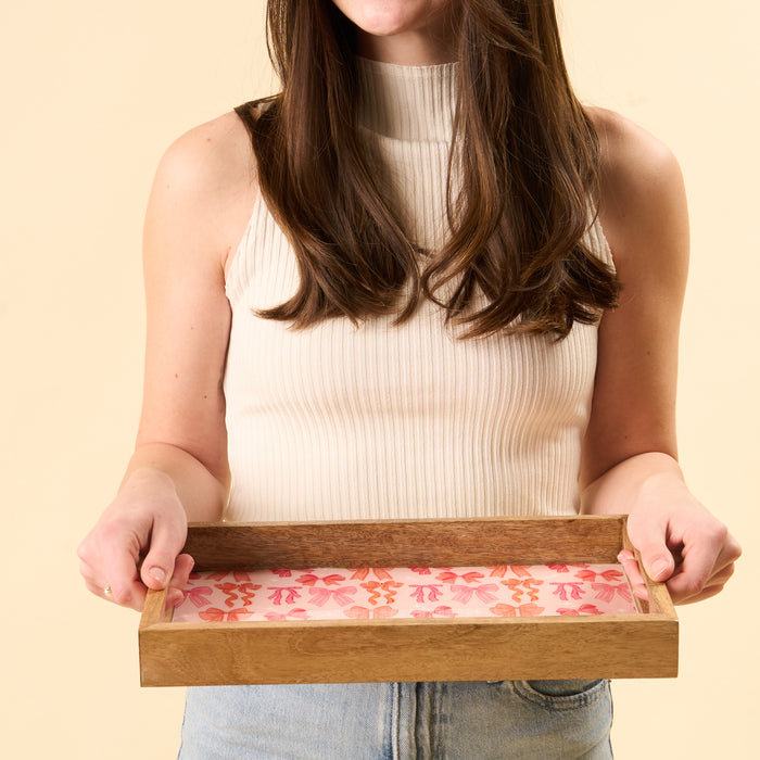 A woman in a sleeveless white top and jeans holds the-darling-effect's Blushing Bows Rectangle Wood Enamel Tray, featuring a pink and red floral design. Her face isn't shown, with a plain light beige background behind her.