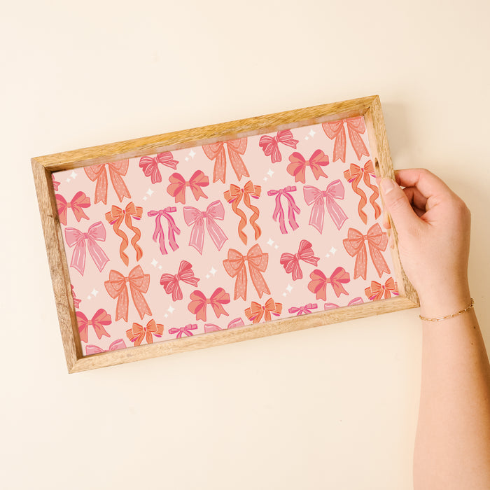 A hand holds the-darling-effect’s Blushing Bows Rectangle Wood Enamel Tray, lined with paper decorated in pink and orange bows on a light pink background with tiny white stars.