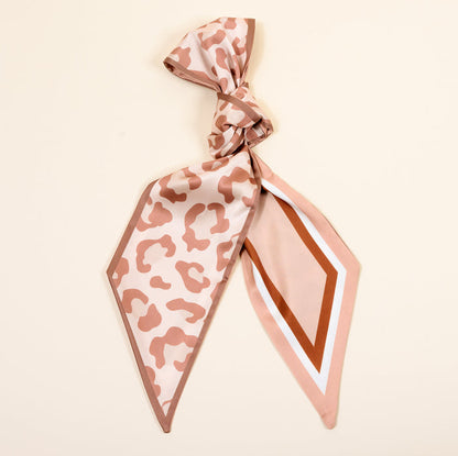 The Darling Effect's Born Wild Twirl & Tie Scarf Accessory features a knotted top, beige and light brown leopard print on one end, and brown, white, and pink geometric stripes on the other for endless styling against a light cream background.