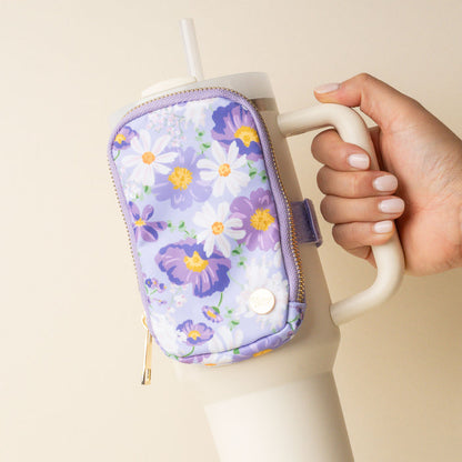 A hand holds a cream tumbler with the-darling-effect's Bouquet Beauty Purple Tumbler Fanny Pack, a water-resistant pouch adorned with white and yellow flowers that doubles as a trendy fanny pack for your drink.