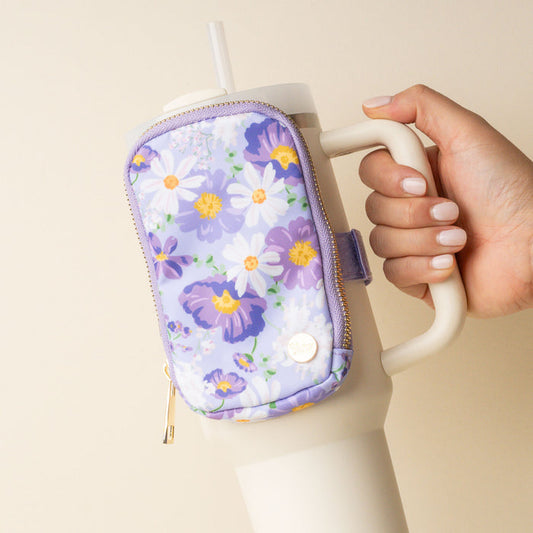 A hand holds a cream tumbler with the-darling-effect's Bouquet Beauty Purple Tumbler Fanny Pack, a water-resistant pouch adorned with white and yellow flowers that doubles as a trendy fanny pack for your drink.