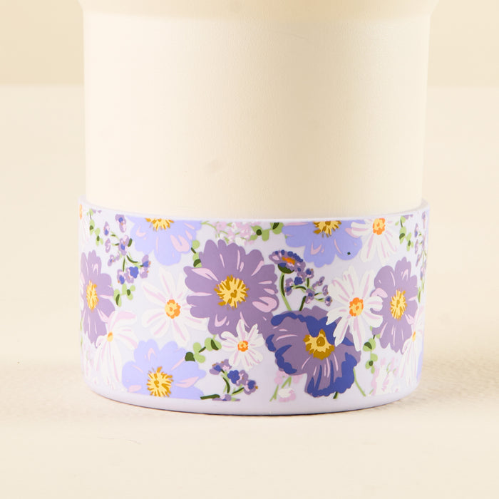 The Bouquet Beauty Purple Tumbler Silicone Boot from the-darling-effect features a white cup adorned with purple, lavender, and white flowers with green leaves around the base and includes a silicone boot for added protection.