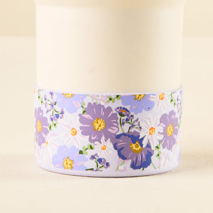 The Bouquet Beauty Purple Tumbler Silicone Boot from the-darling-effect features a white cup adorned with purple, lavender, and white flowers with green leaves around the base and includes a silicone boot for added protection.