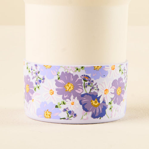 The Bouquet Beauty Purple Tumbler Silicone Boot from the-darling-effect features a white cup adorned with purple, lavender, and white flowers with green leaves around the base and includes a silicone boot for added protection.