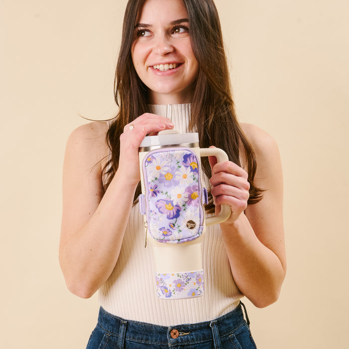 A smiling woman in a sleeveless beige top and blue jeans holds the-darling-effect’s Bouquet Beauty Purple Tumbler Silicone Boot with a matching floral pouch, standing against a plain light background.