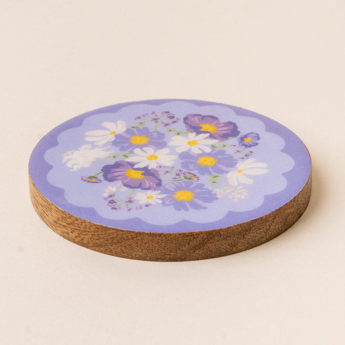 The-darling-effect's Bouquet Beauty Purple Wood Enamel Coaster features a round design with a purple top, adorned with white and purple flowers. This charming coaster protects surfaces while adding a decorative touch.