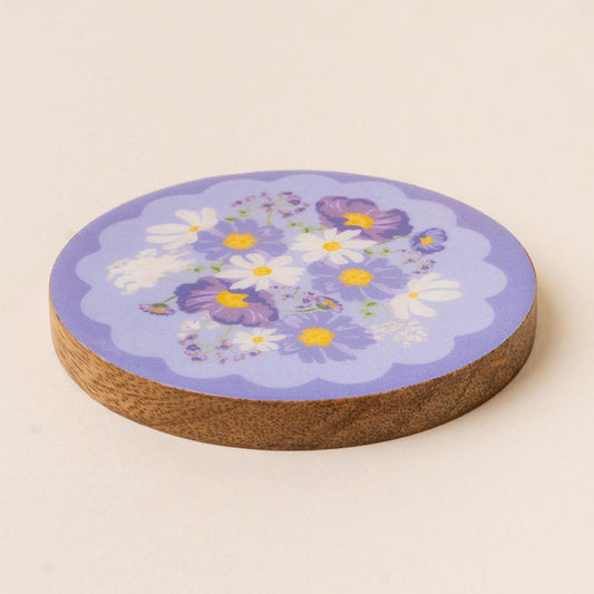 The-darling-effect's Bouquet Beauty Purple Wood Enamel Coaster features a round design with a purple top, adorned with white and purple flowers. This charming coaster protects surfaces while adding a decorative touch.