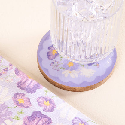 A glass of ice water rests on the-darling-effect’s Bouquet Beauty Purple Wood Enamel Coaster, featuring a floral design. A matching cloth beside it adds charm—perfect for those who love stylish and durable coasters.