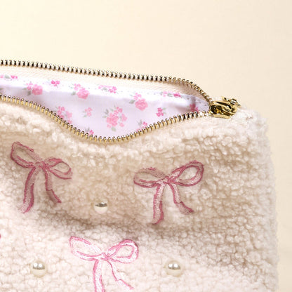 The Bows Pink Teddy Pouch by the-darling-effect features cream sherpa fabric, embroidered pink bows, pearl accents, and a white lining with a pink floral design—perfect for storing accessories or teddy essentials.