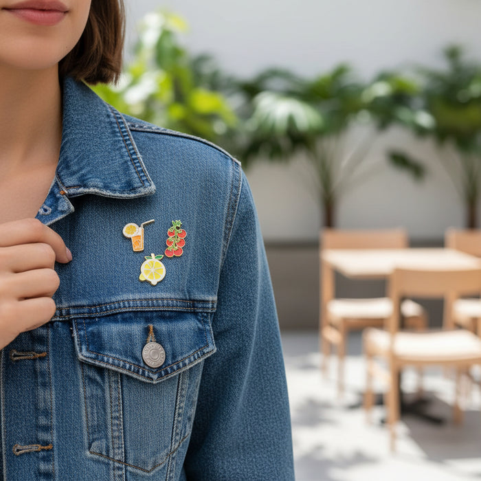 A person wears The Darling Effect’s Capri Cutie The Pin Pack—expressive pins shaped like a drink, lemon slice, and cherries—on a blue denim jacket while standing outdoors among blurred chairs, tables, and green plants.