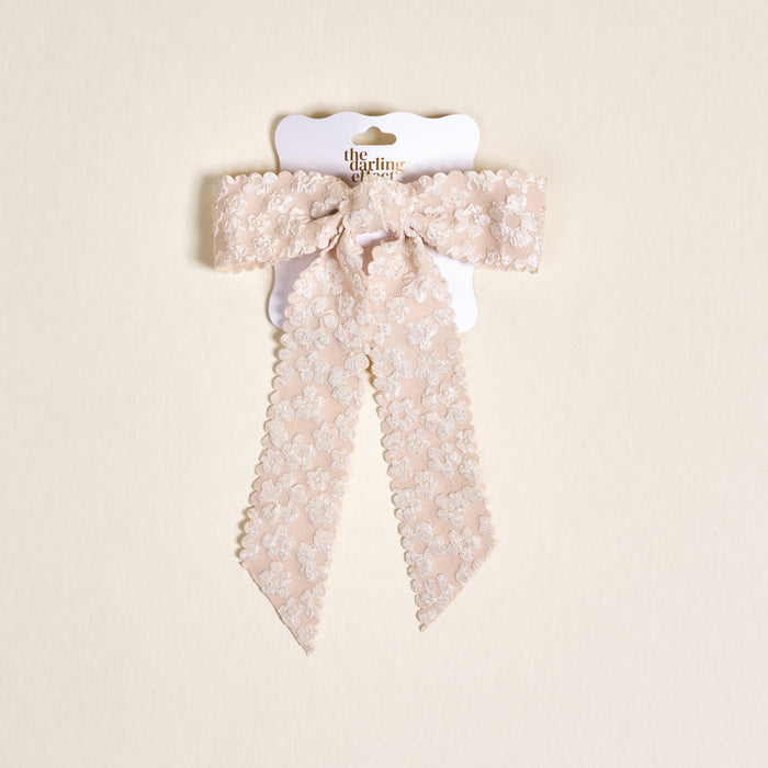 The Champagne Flower Scallop Hair Bow by the-darling-effect features pale pink lace with a floral pattern and scalloped edges, clipped to a white card with gold text on a beige background—an elegant addition to your hair accessory collection.