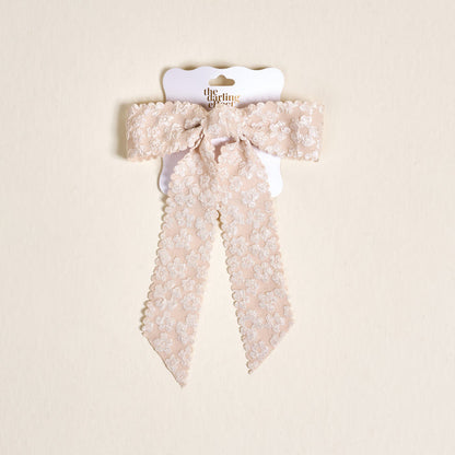 The Champagne Flower Scallop Hair Bow by the-darling-effect features pale pink lace with a floral pattern and scalloped edges, clipped to a white card with gold text on a beige background—an elegant addition to your hair accessory collection.