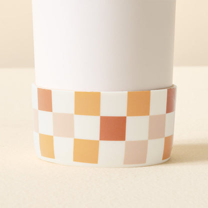 A close-up of The Darling Effect Check Brown Tumbler Silicone Boot, a white cylindrical tumbler accessory with a checkered beige, orange, brown, and white design, shown on a light surface for extra tumbler protection.