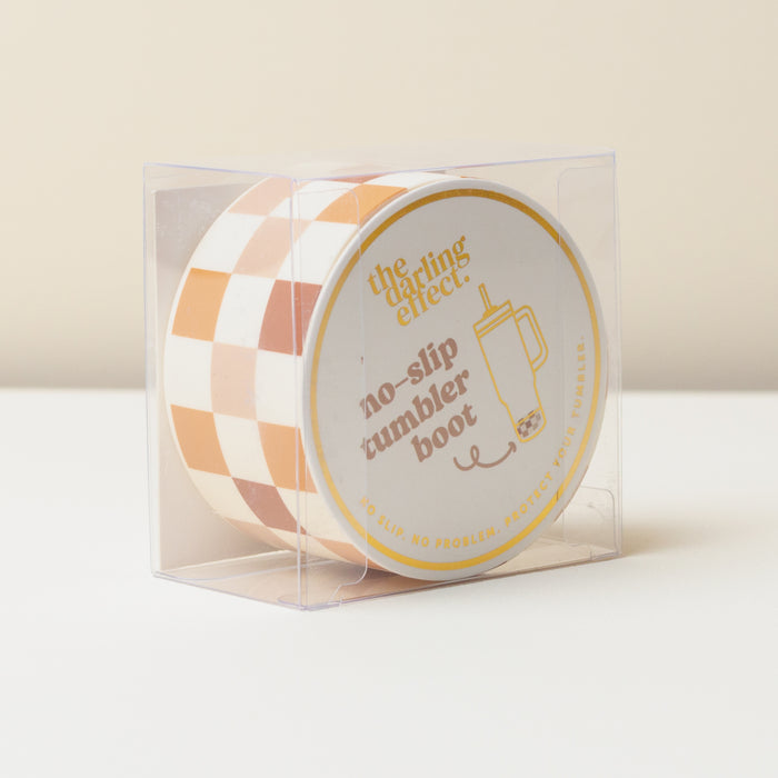 The Darling Effect Check Brown Tumbler Silicone Boot protects your tumbler in style. Displayed in a clear plastic box, its label reads "No-slip tumbler boot"—a must-have accessory for every tumbler owner.