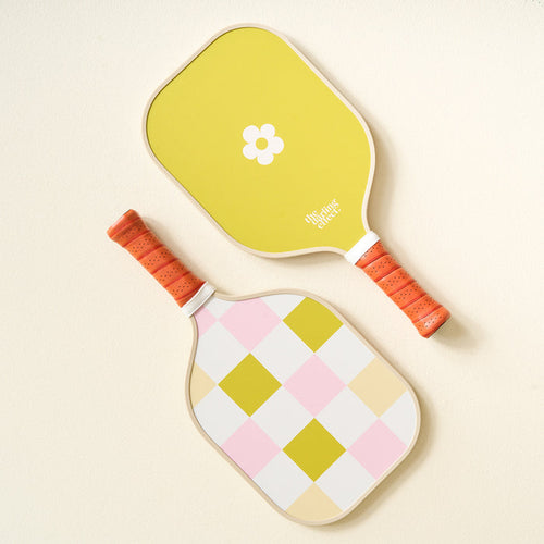 Two top-quality pickleball paddles by The Darling Effect: one with a white flower on yellow, the other is the Check Green Pickleball Paddle with a pastel checkered pattern. Both have orange handles and are set against a light cream background.