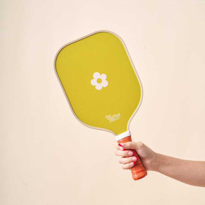 A hand with pink nail polish holds The Darling Effect’s Check Green Pickleball Paddle, featuring a unique white flower design in the center and an orange handle, set against a plain light background.
