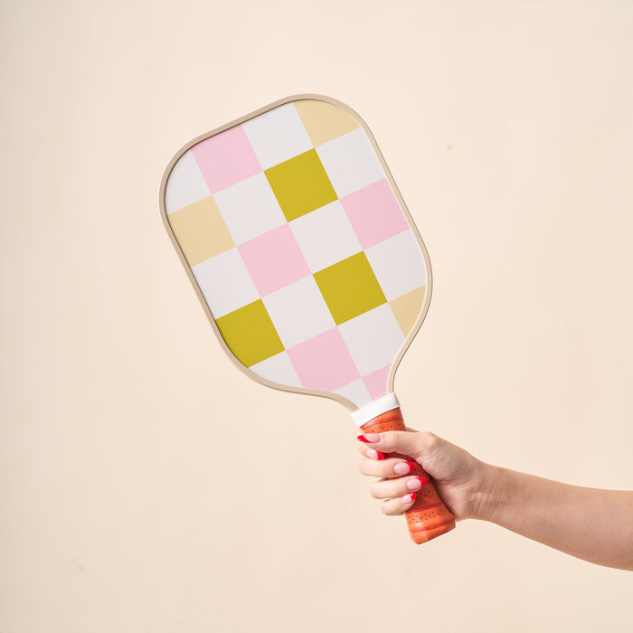 A hand with pink-painted nails holds The Darling Effect's Check Green Pickleball Paddle, featuring a unique green checkered design and a brown leather grip, against a plain beige background.