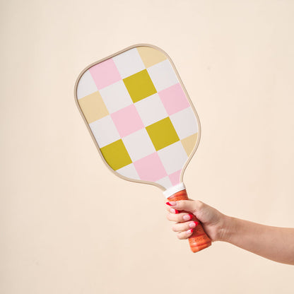 A hand with pink-painted nails holds The Darling Effect's Check Green Pickleball Paddle, featuring a unique green checkered design and a brown leather grip, against a plain beige background.