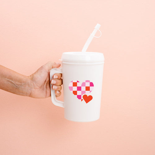 A hand holds the-darling-effect's Checkered Heart 22oz Mega Mug, a white insulated cup with handle, lid, and straw. It features a large pink checkered heart and a small red heart on a light peach background.