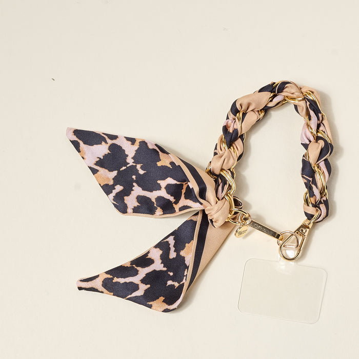 The Darling Effect’s Cheetah Call Me Later Phone Scarf features tan and black leopard-print fabric, gold-tone chain links, a matching bow, gold clasp, and a clear rectangular tag—an ultra-chic accessory for your phone.
