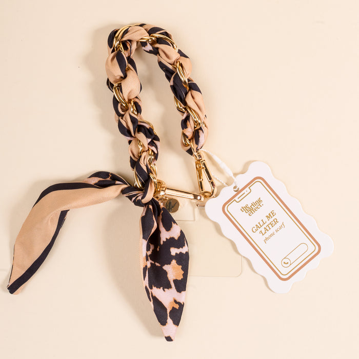 The Darling Effect’s Cheetah Call Me Later Phone Scarf features a gold-tone chain with beige and black cheetah-print fabric tied in a bow, plus a “CALL ME LATER Phonegrip” tag—the perfect stylish accessory for your device.