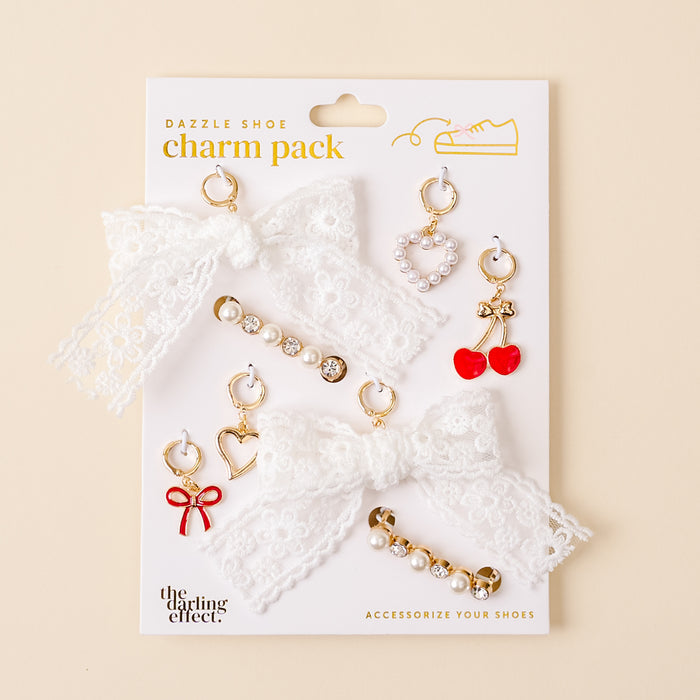 The Cherry Twist Dazzle Shoe Charm Pack by the-darling-effect features two white lace bows, gold and pearl sneaker charms, red cherry and heart shoe charms, and rhinestone accents on a card.
