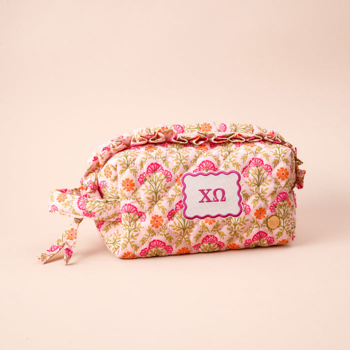 The Darling Effect’s Chi Omega Essentials Quilted Cosmetic Pouch features a pink, orange, and green floral design and a purple ΧΩ patch—perfect for Sorority Essentials. Shown against a light beige background.