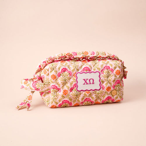 The Darling Effect’s Chi Omega Essentials Quilted Cosmetic Pouch features a pink, orange, and green floral design and a purple ΧΩ patch—perfect for Sorority Essentials. Shown against a light beige background.