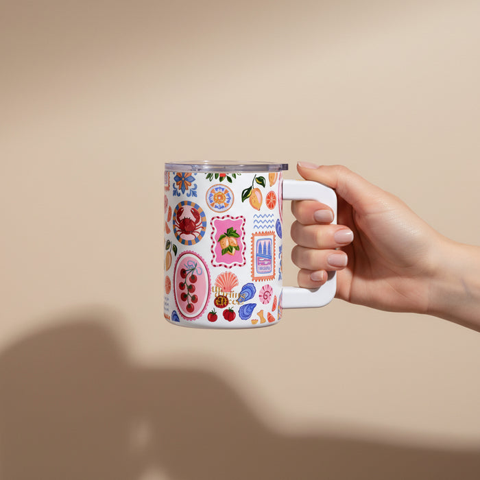 A hand holds The Darling Effect’s Ciao Bella 14 oz Insulated Mug, featuring colorful fruit and floral illustrations with decorative frames, set against a plain beige background.